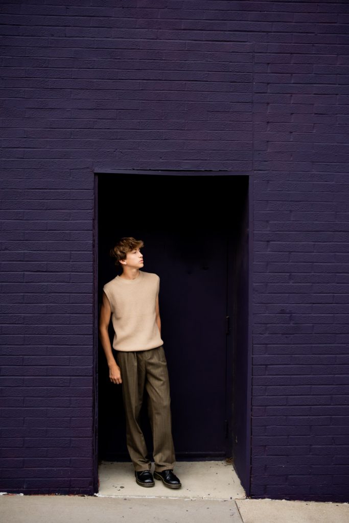 Senior boy standing in a purple doorway against a brick wall.