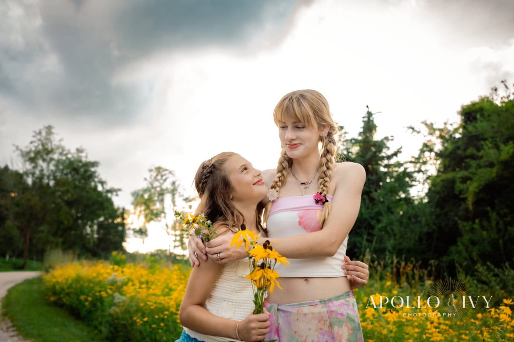 Best timing for family pictures: A month-by-month guide Older sister and younger sister embracing in front of a field of yellow flowers in Iowa