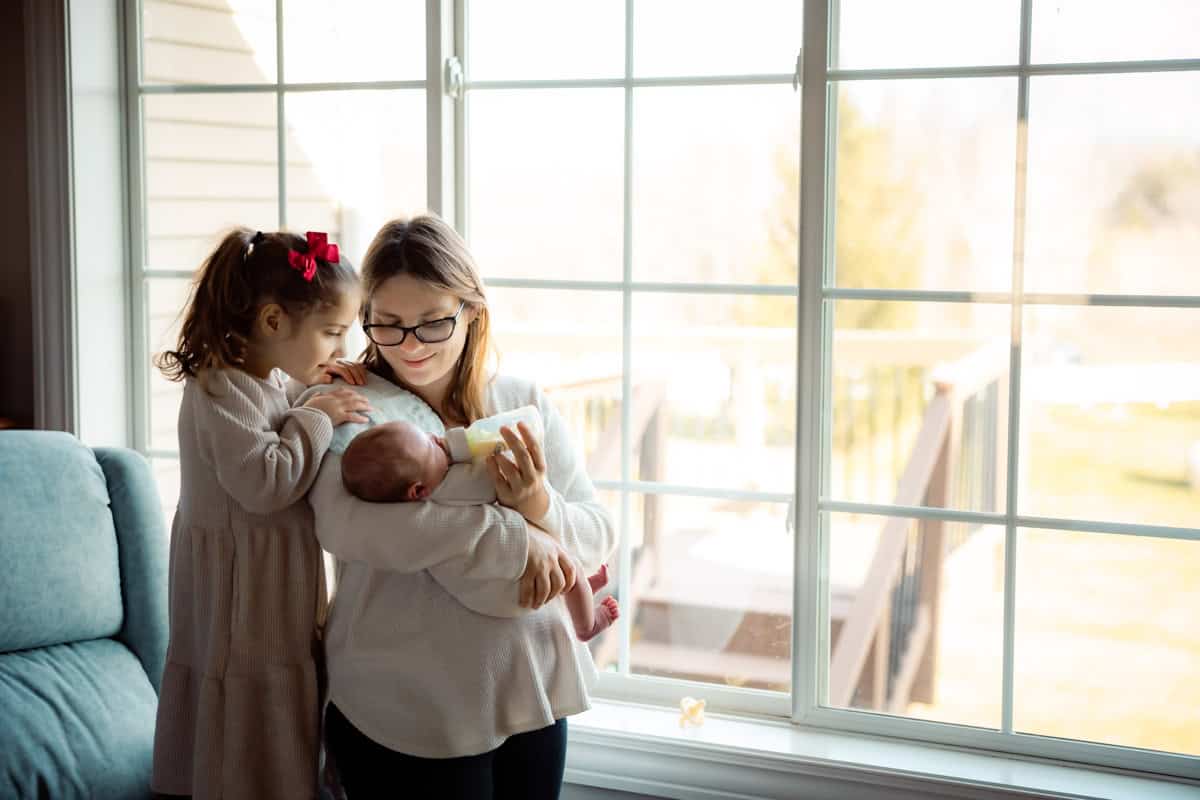 Mom, toddler girl and newborn by a window