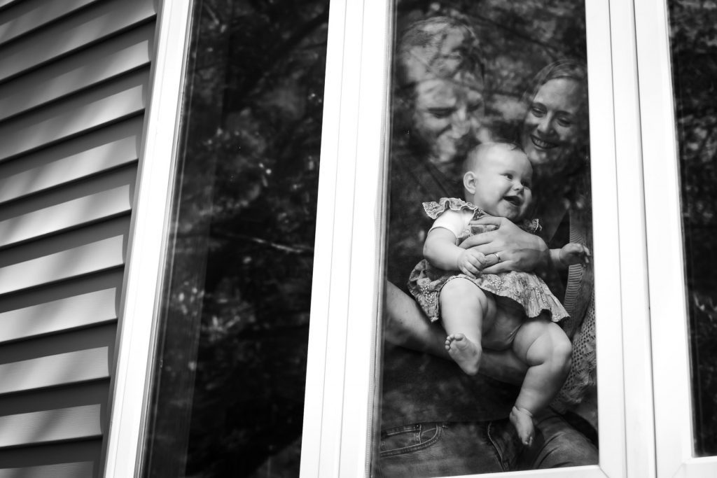 Mom and dad holding a squishy baby next to a window with photo taken from outside the house.