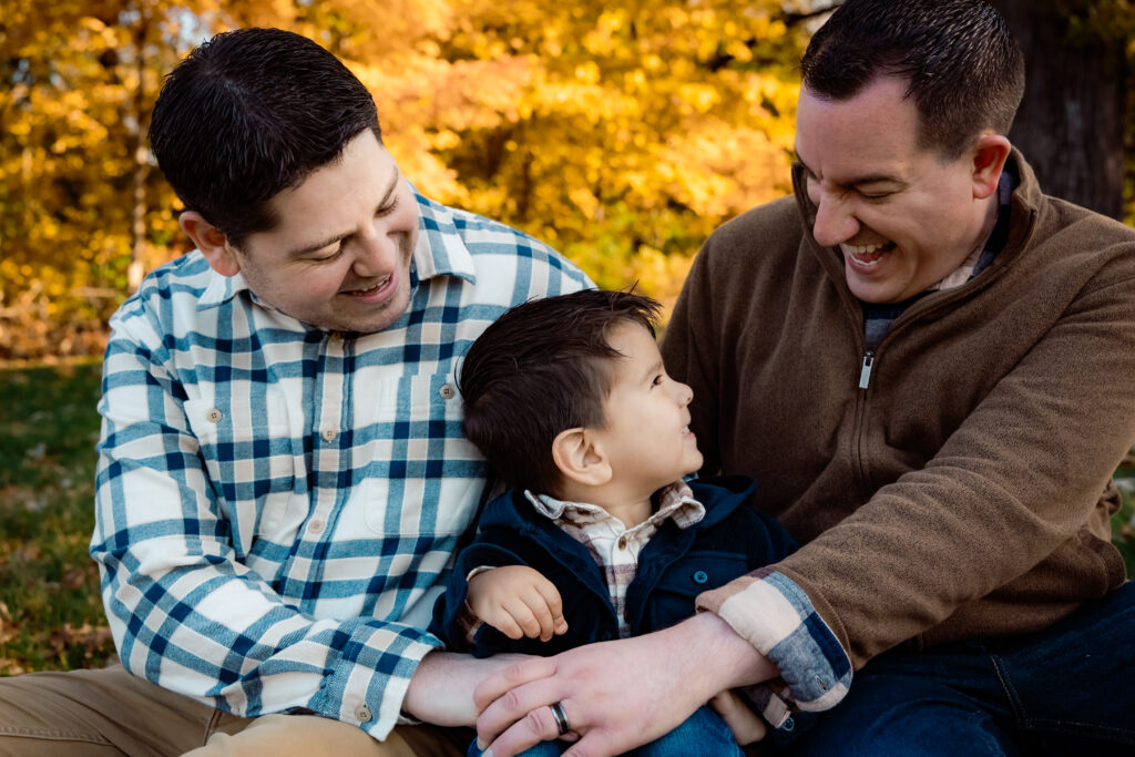 What to wear for cold-weather outdoor photos Photo of a family of three where one dad is wearing a plaid shirt and the other dad is wearing a button down under a dark brown quarter zip. The toddler boy between them is wearing a plaid button down under a navy blue button down corduroy shacket.