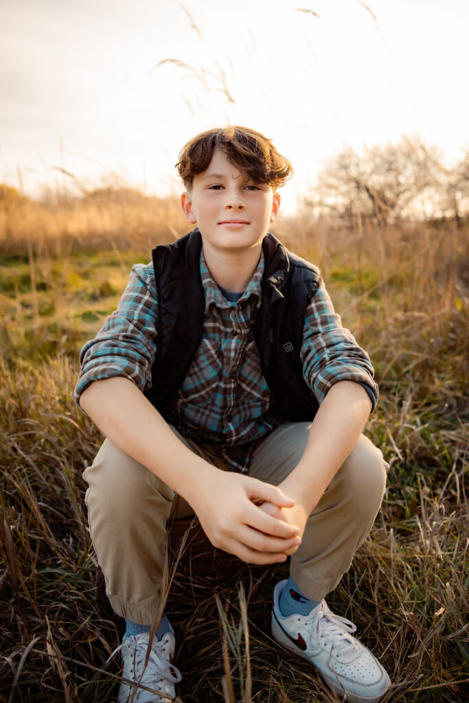 What to wear for cold-weather outdoor photos Young teen wearing a flannel shirt with a black light pile vest over top, seated in a grassy field.