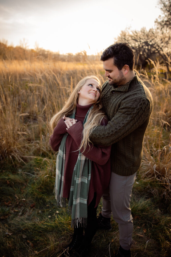 What to wear for cold-weather outdoor photos Photo of a woman in sweater wearing a scarf cuddles with her husband in a grassy field in the fall.