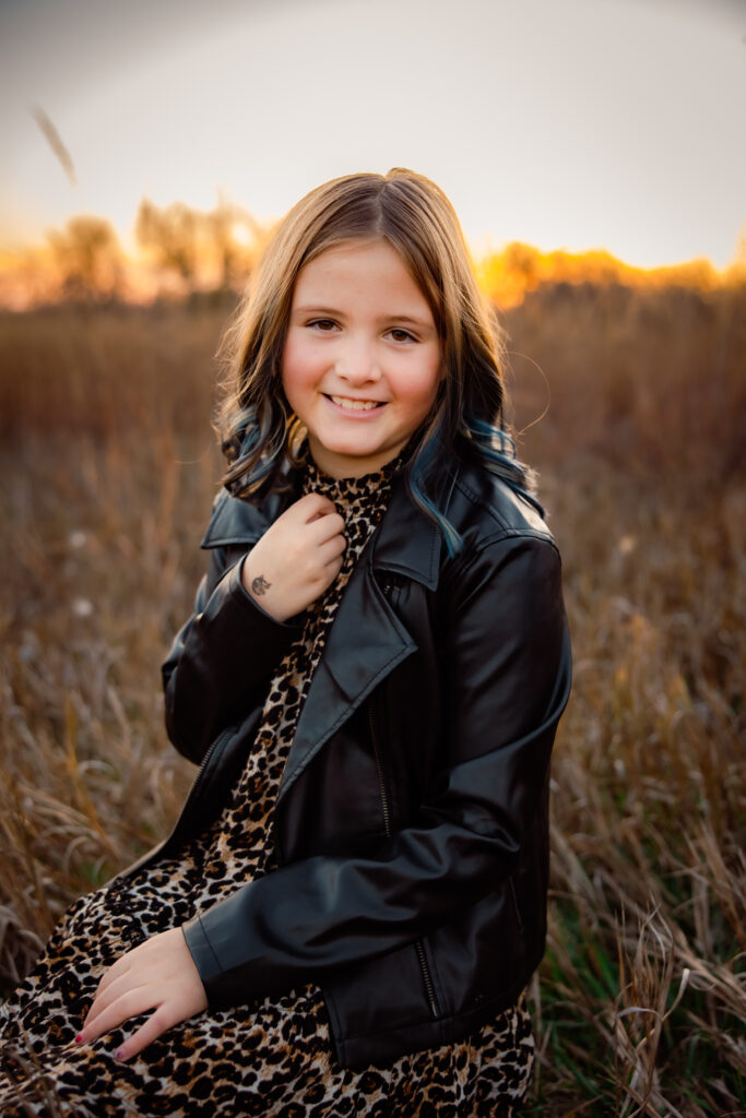 What to wear for cold-weather outdoor photos Photo of young girl sitting in a grassy field at sunset wearing an animal print dress and black leather jacket, smiling at the camera.