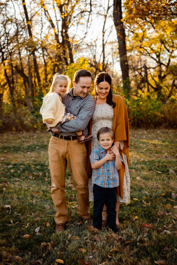 What to wear for cold-weather outdoor photos Photo of a family in the fall, standing in grass covered with leaves and trees in the background. Children are toddler age, with a girl in dad's hands and a little boy in front who appears to be quite cold in short sleeves.