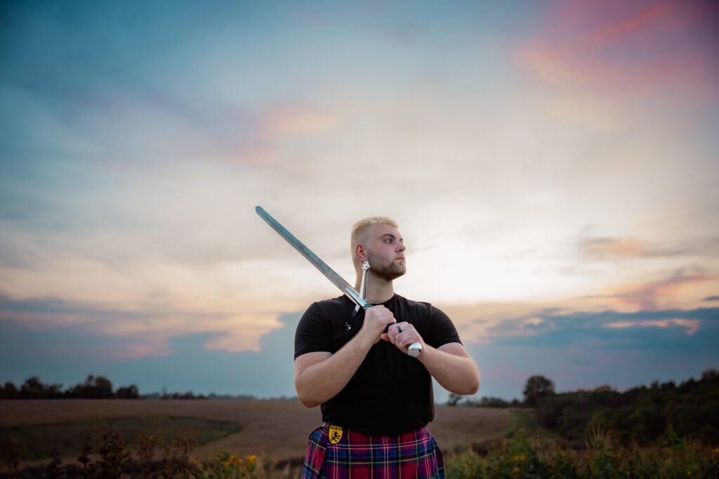 Beginner's guide to senior pictures in Iowa Senior guy in a black tee and kilt holds a sword over one shoulder while looking away against a sunset sky