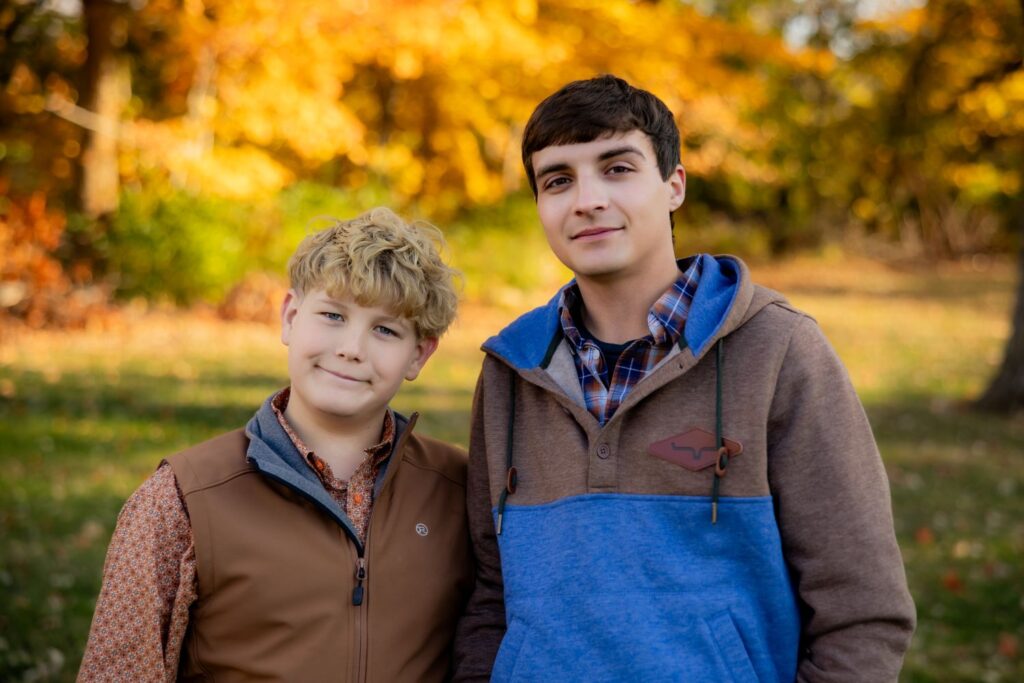 Best colors to wear for photos in the fall Two brothers stand side by side looking at the camera and slightly smiling with a fall backdrop. The teen on the right is wearing a brown and light blue jacket over a button down.