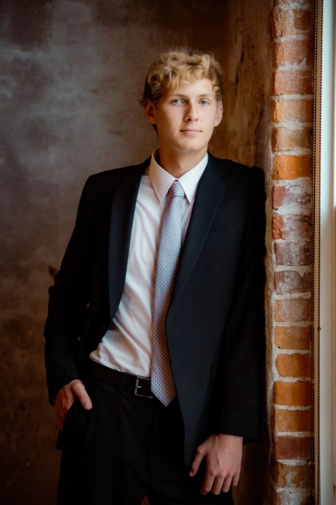 Senior boy in a suit leaning against exposed brick wall
