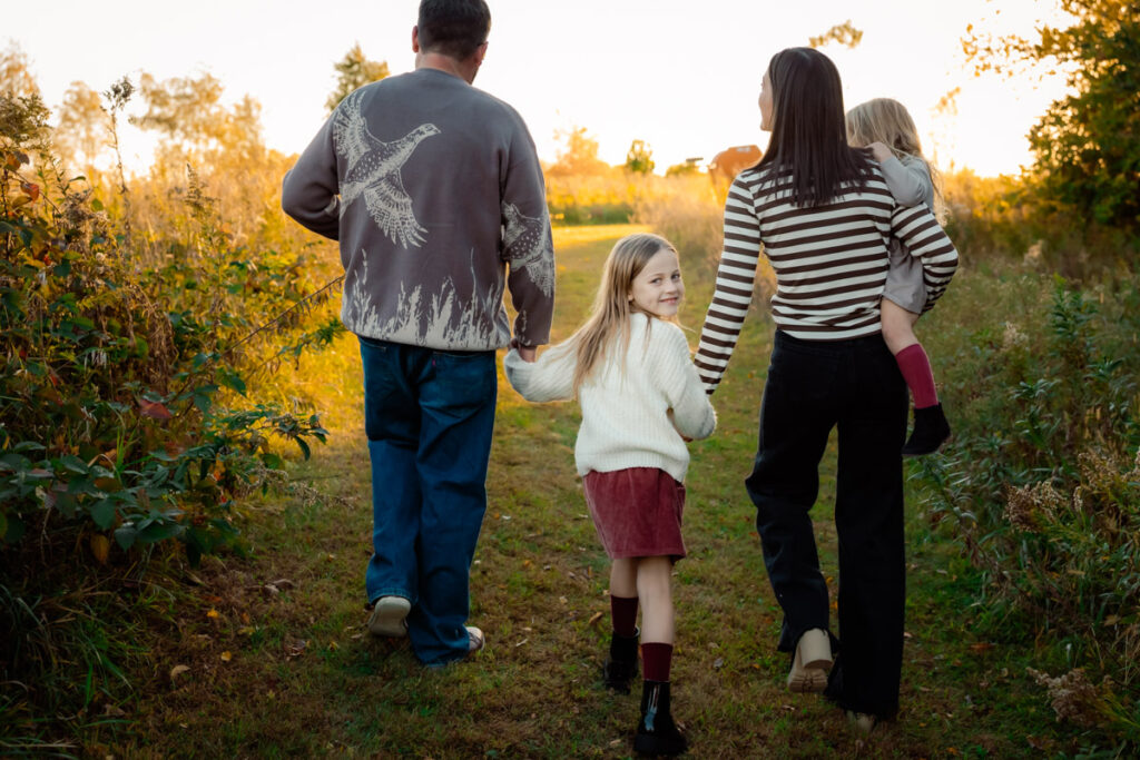 Family of four walking away from the photographer with a little girl around six turning back to smile at the camera