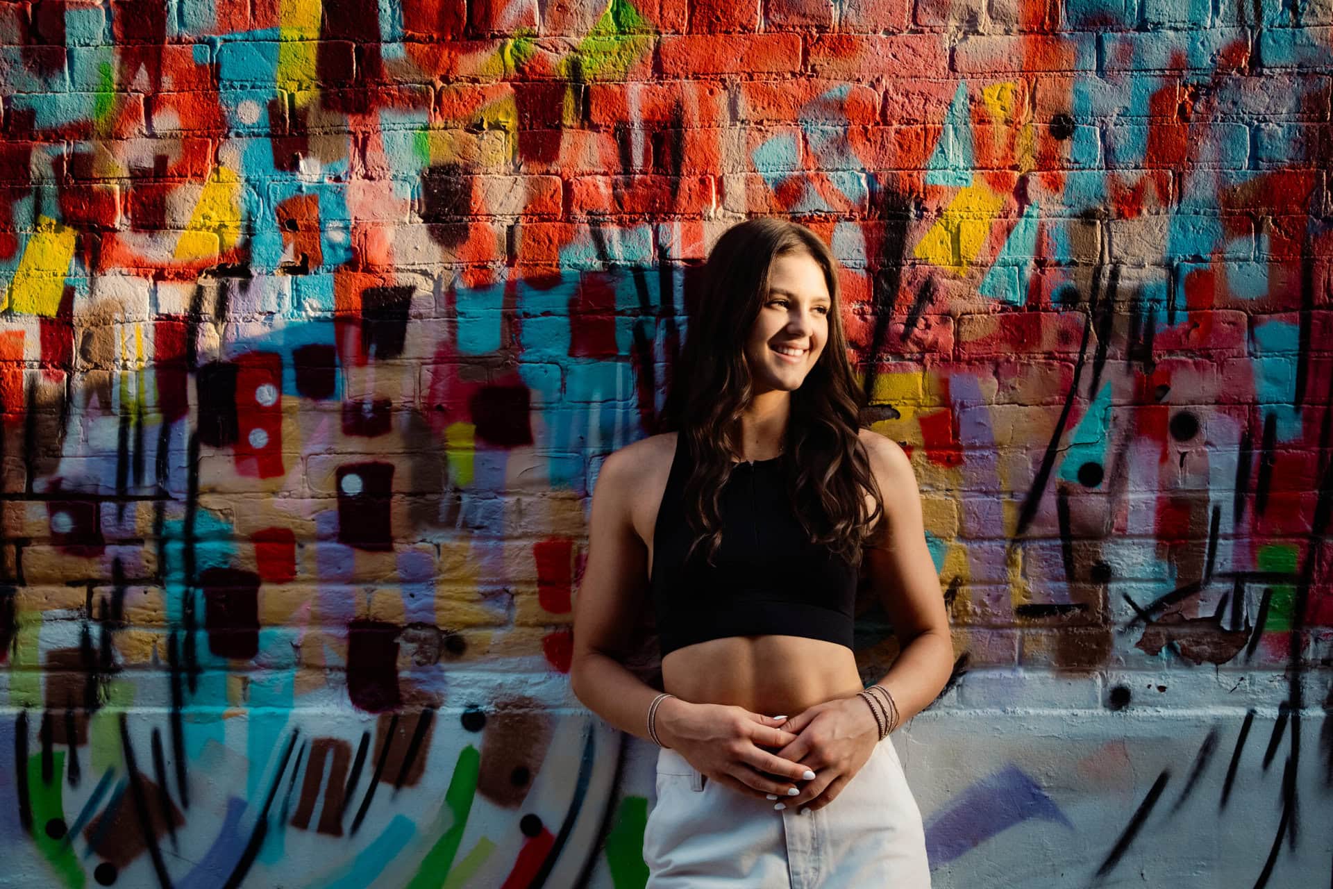 Senior girl leaning against colorful mural brick wall looking into setting sun