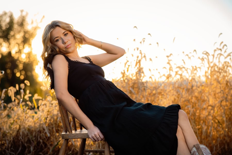 Senior girl in black dress leaned back on a wooden chair in front of a sunset