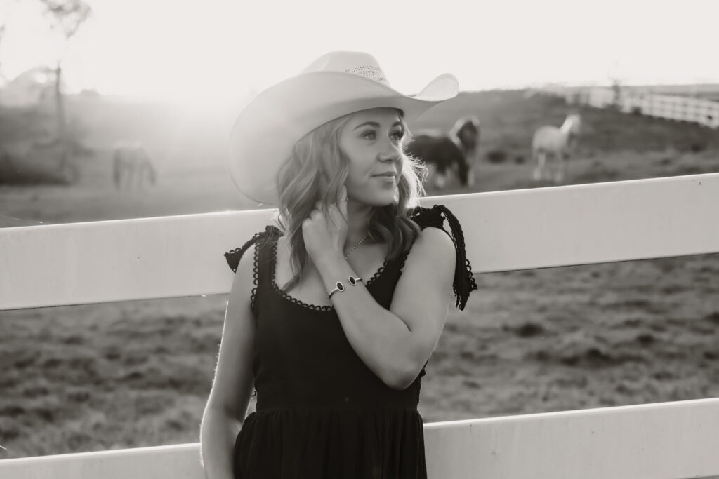 Beginner's guide to senior pictures in Iowa Senior girl next to white vinyl fence in black dress wearing a cowboy hat with horses in the background
