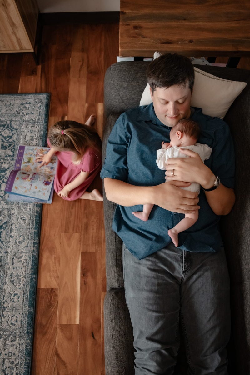 Father lies reclined on a couch with a newborn sleeping on his chest while toddler girl sits on the floor with her back to the couch reading a book