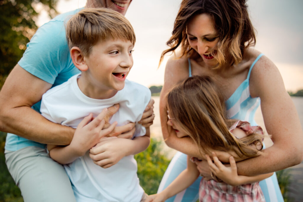 Should I wear my hair down for family photos? Family of four in a chaotic embrace with mom and dad sneaking up behind the kids to hug them on a windy day by the lake.