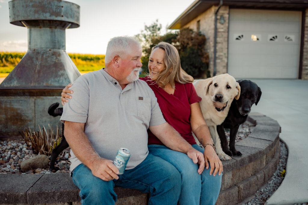 Should I wear my hair down for family photos? Man and woman sitting in front of their home with two labradors on a windy fall afternoon while the man holds a Busch Light