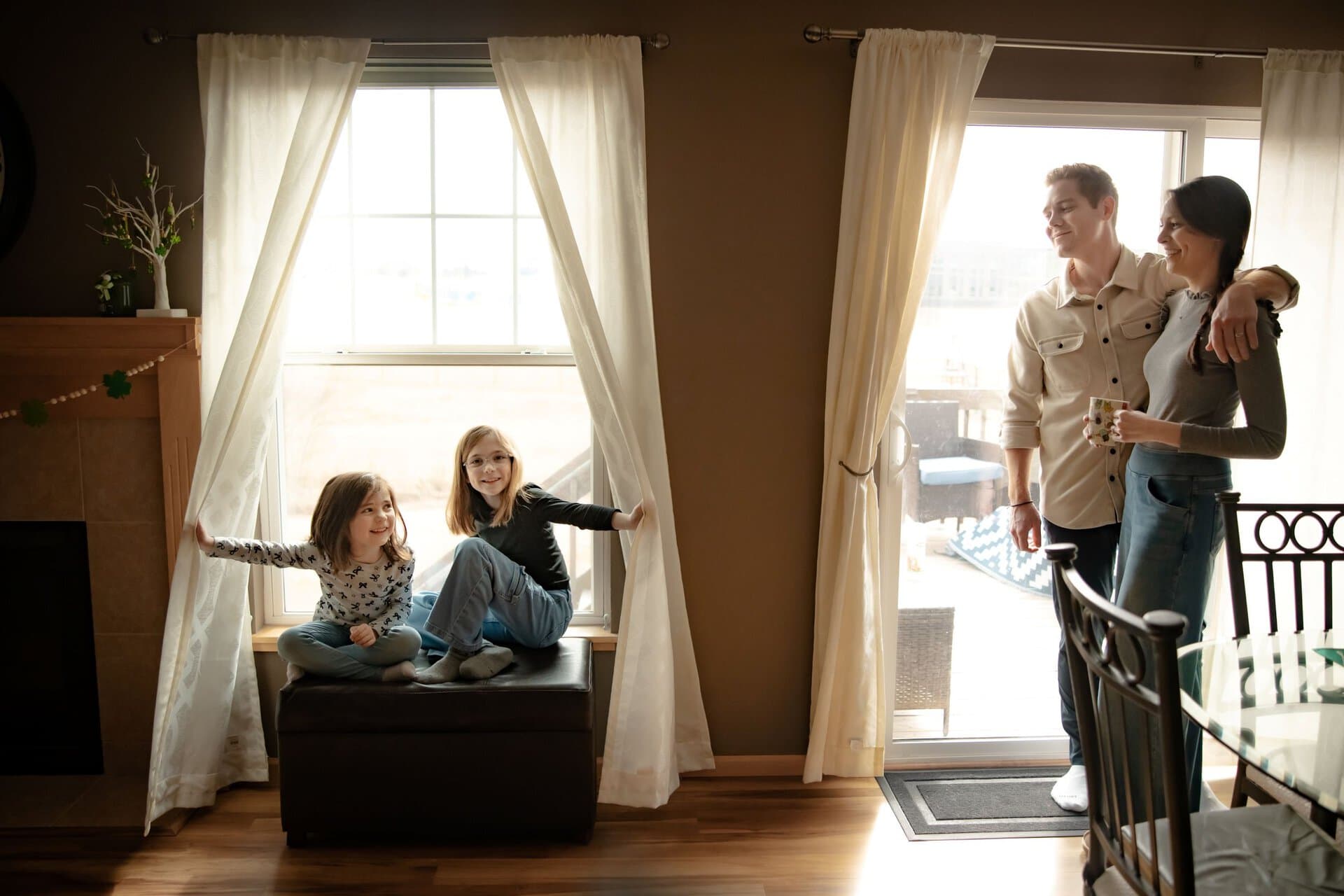 A family of four in their living room with mom and dad standing in front of the sliding glass door looking at their two daughters seated nearby who are looking back and smiling