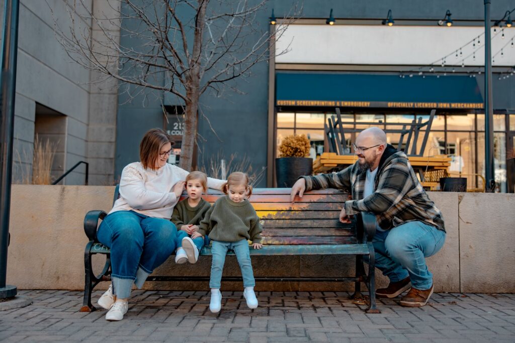Best timing for family pictures: A month-by-month guide Parents sitting on a bench in downtown Iowa City area while daughters sit next to mom who is at the left end and dad squatting next to the right end.