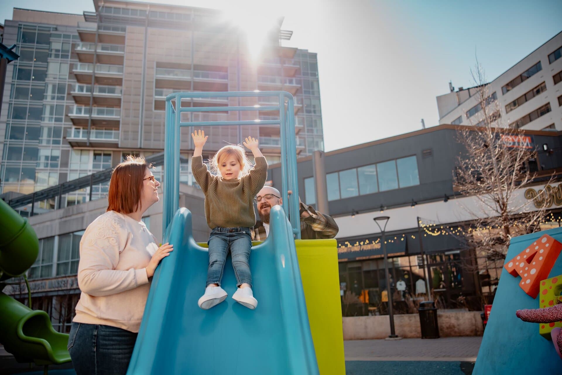 Best timing for family pictures: A month-by-month guide Parents on playground in downtown Iowa City area watching toddler daughter go down the slide with her hands in the air