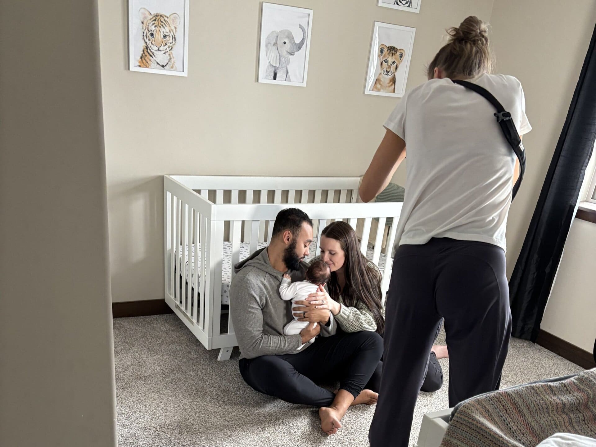 Behind the scenes at a newborn photo shoot with photographer standing back to camera photographing a couple holding a newborn sitting on the floor up against the crib