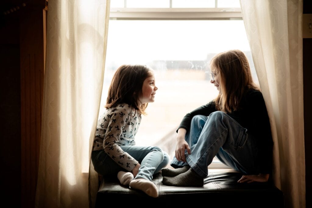 Two young girls sit on a cushioned ottoman in front of a window and smile at each other