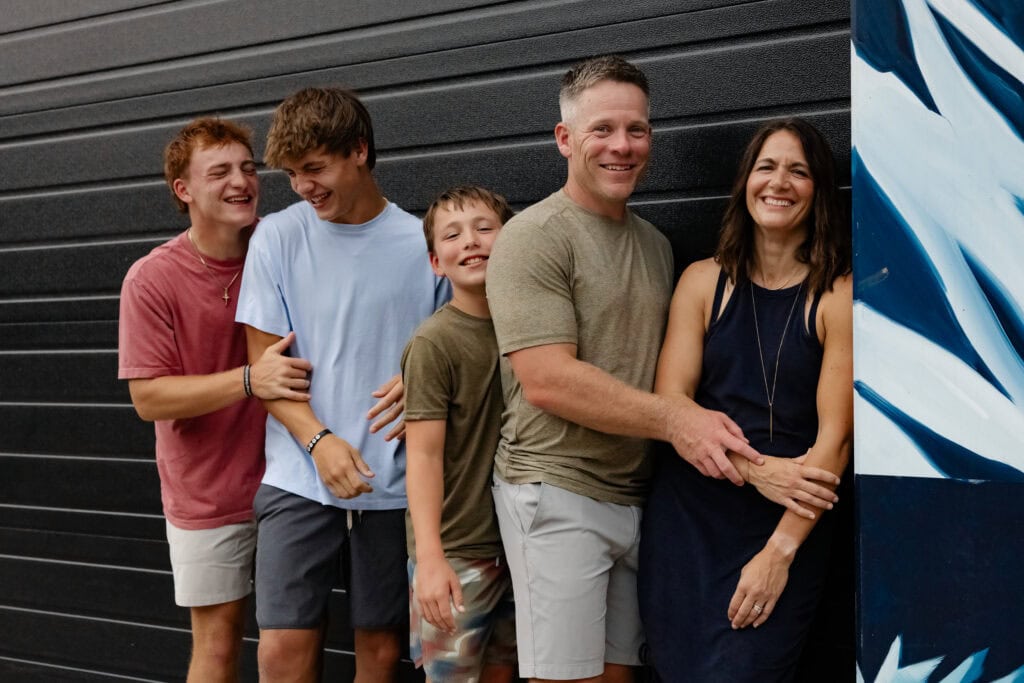 Family of five with three boys standing in a row, with mom, dad, and youngest son looking at camera while older 2 giggle maniacally.