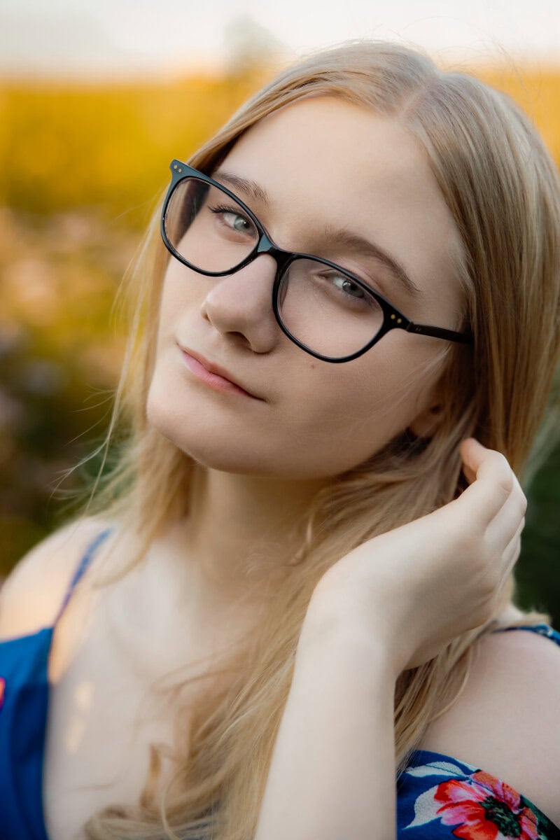 Senior girl in a dress in a flower field with long blonde hair and glasses tucking her hair behind her ear