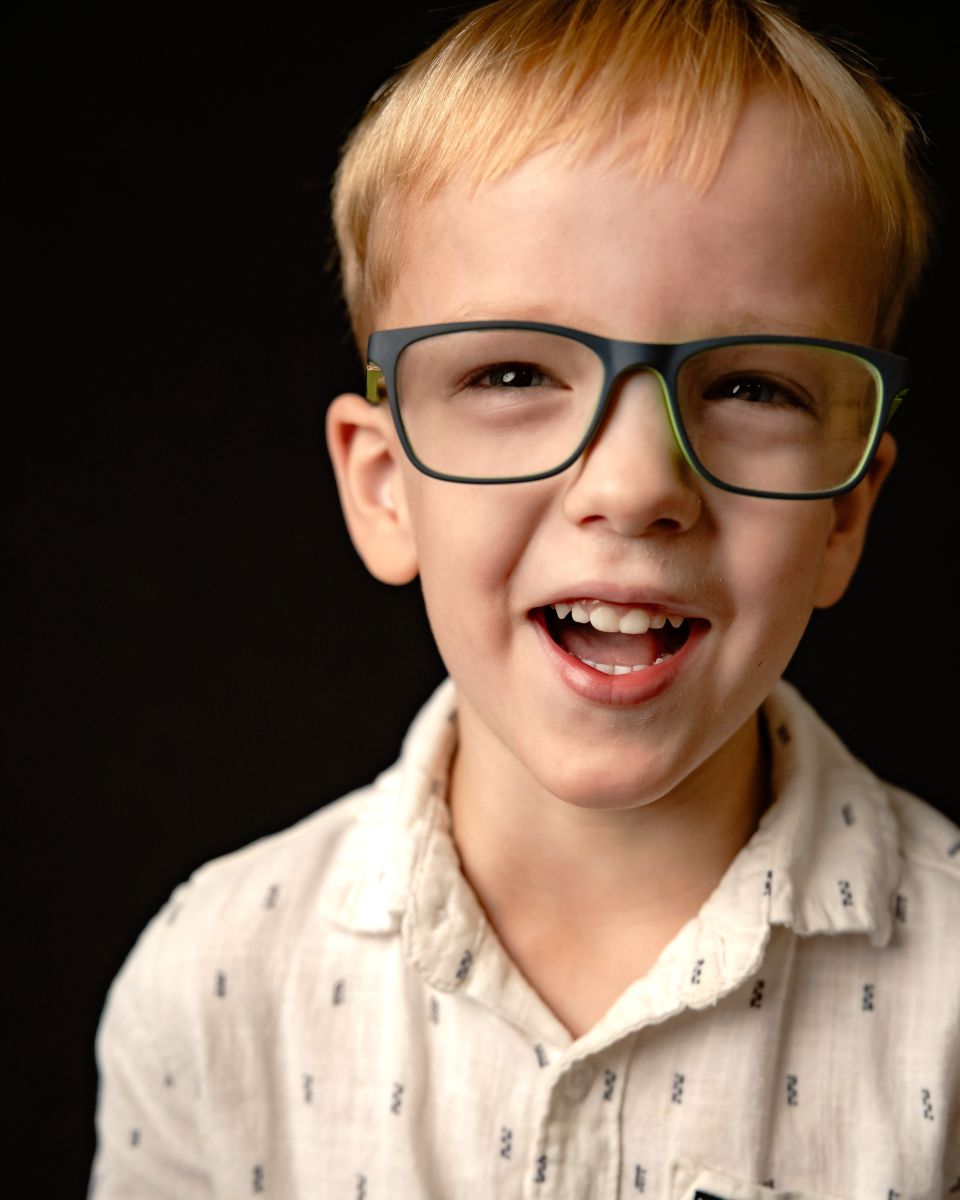 Toddler boy with blonde hair, glasses, in a button up shirt laughing in an up close photograph against a black background.