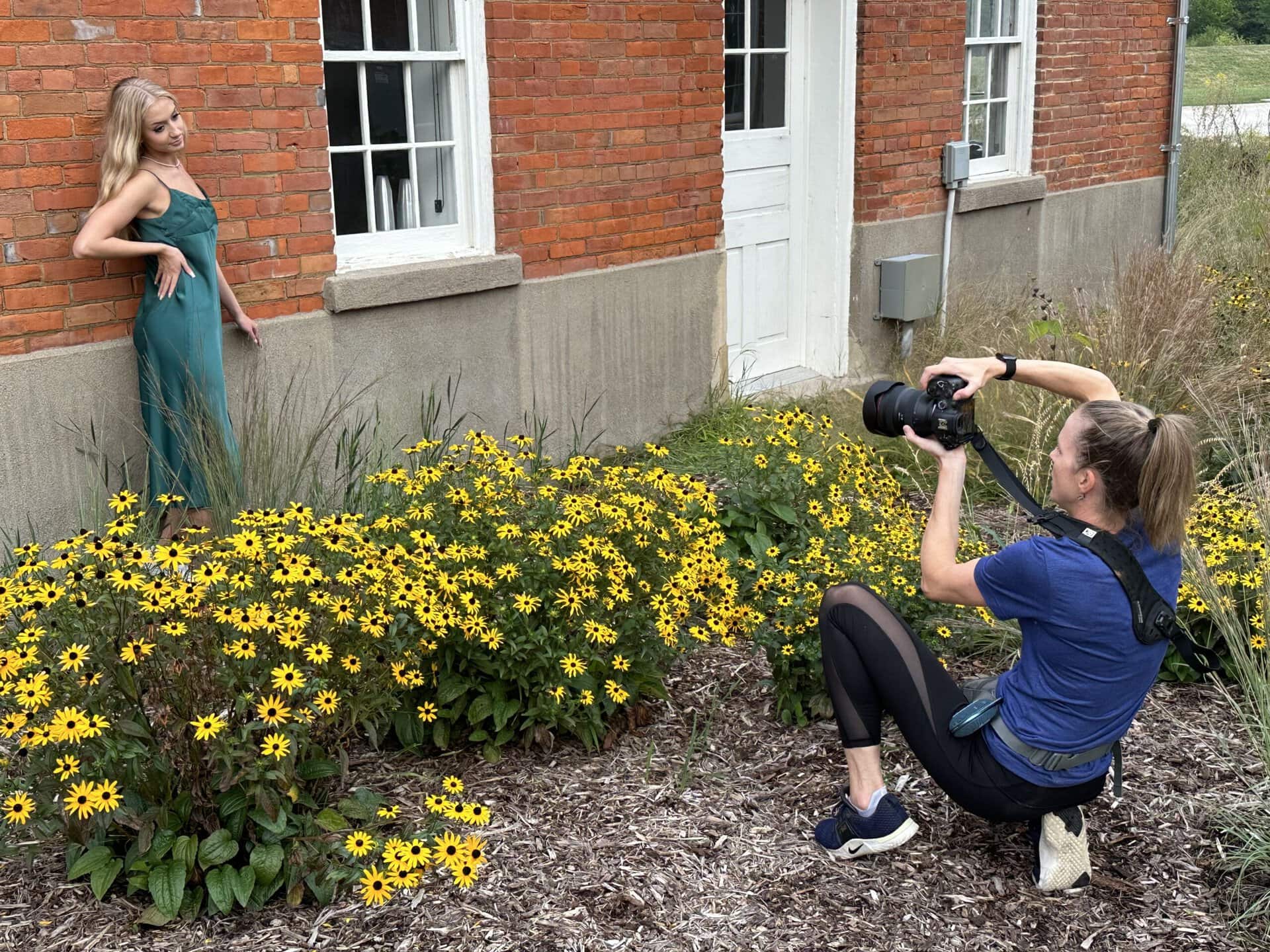 Why I love my SpiderPro v3 Camera Belt System Photographer squatting in front of a row of flowers photographing a senior girl up against a brick wall